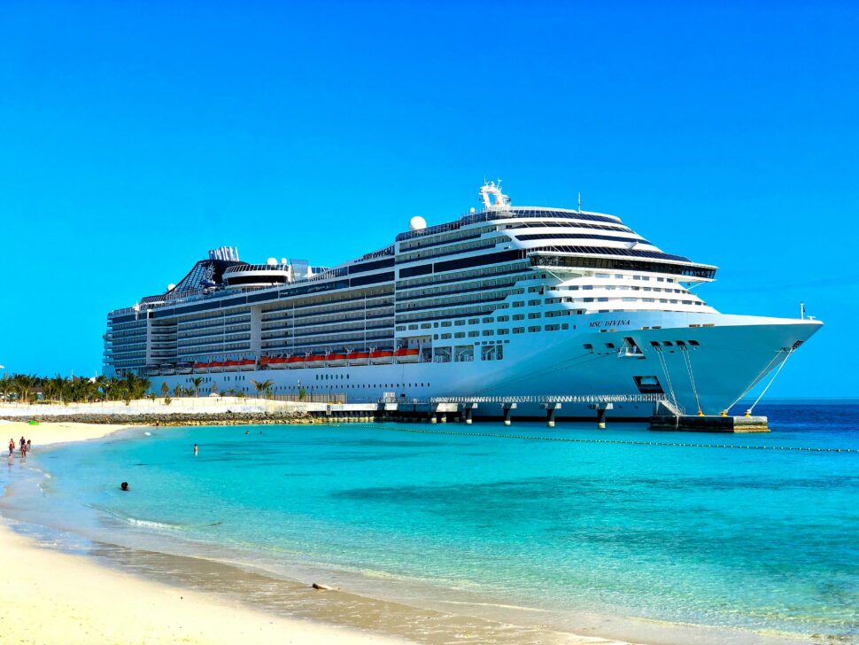 A modern cruise ship docked at a tropical beach port surrounded by lush green trees.