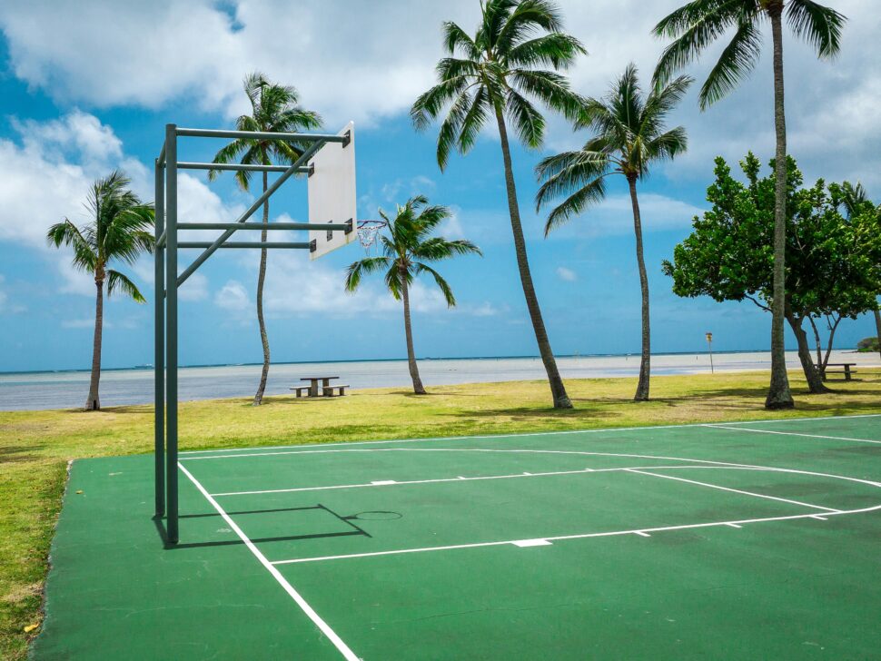 A basketball court in Honolulu, Hawaii, surrounded by palm trees, a beach, and lush grass, showcasing a perfect warm-weather destination.