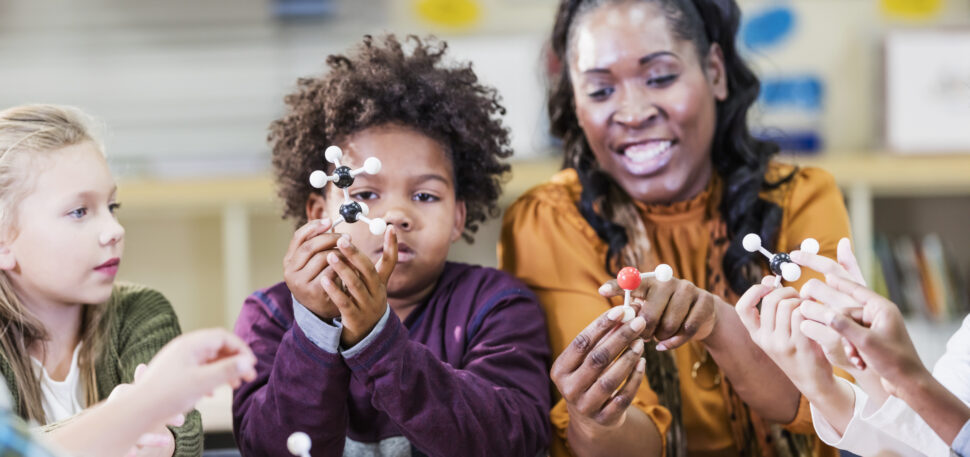 African-American woman teaching a STEM lesson to an elementary school class using models of molecules. She is sitting at a table next to an 8 year old African-American boy who is examining the model he is holding. A little girl is watching. The focus is on the hands and models.