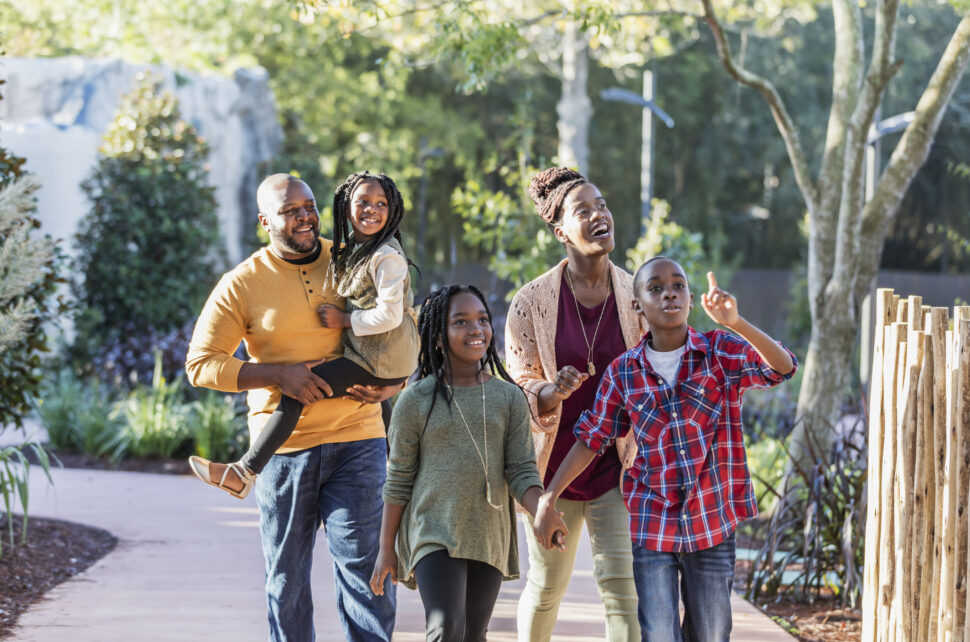 An African-American family with three children visiting a park, walking along a pedestrian walkway surrounded by lush foliate. 