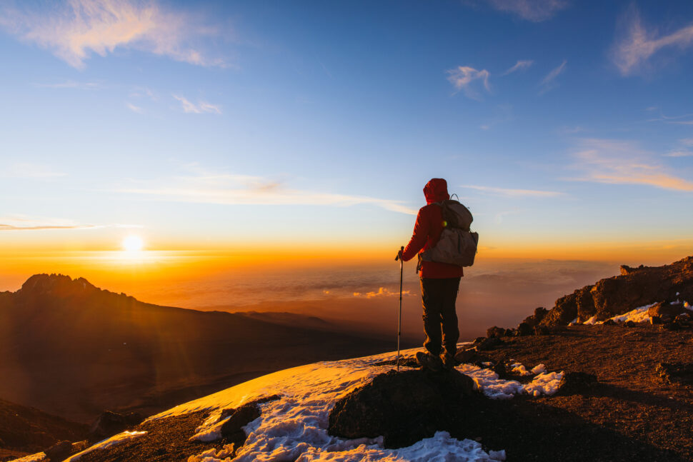 Man with backpack and hiking poles got to the top of Africa - Mount Kilimanjaro and looking at the beautiful bright sunny sunrise above the Meru mountain.