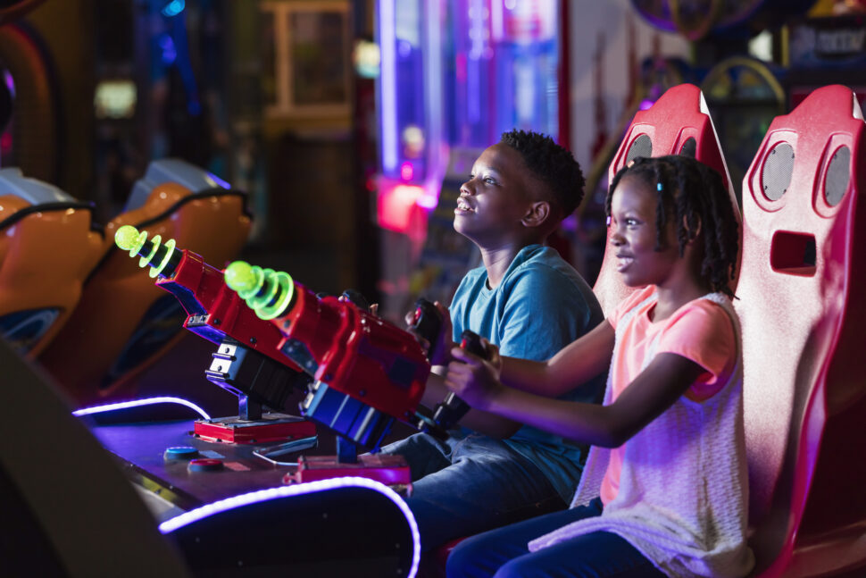 An African-American boy and his sister having fun at an amusement arcade, playing a game pointing toys laser guns at a screen, competing against each other. The main focus is on the boy, 11 years old. The girl is 7.