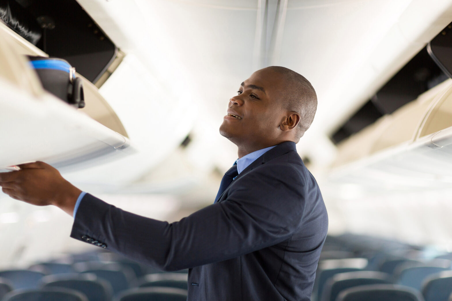 Young businessman putting luggage into overhead bin on airplane.