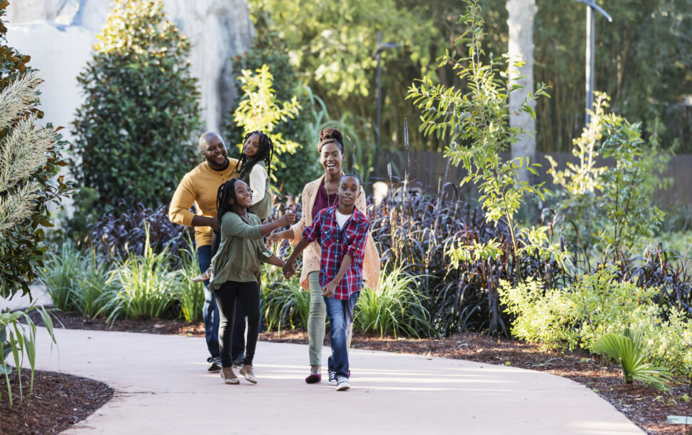 African-American family with three children visiting a park, walking along a pedestrian walkway surrounded by lush foliate.