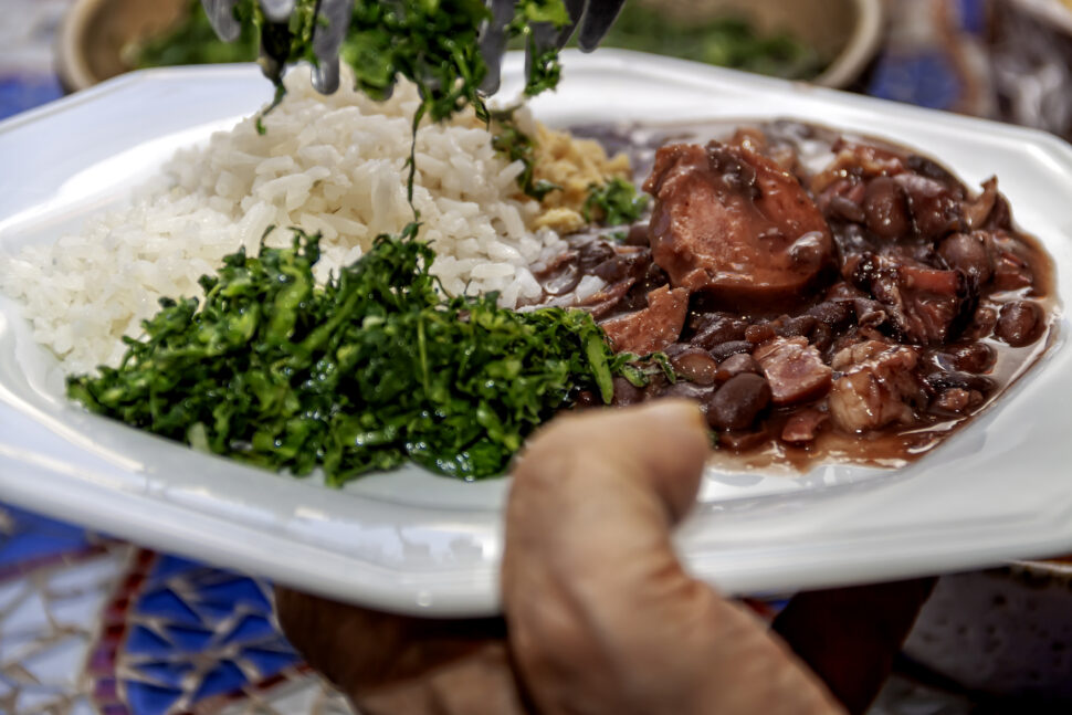 On the plate, the feijoada, white rice , farofa are served and chopped kale is being added.