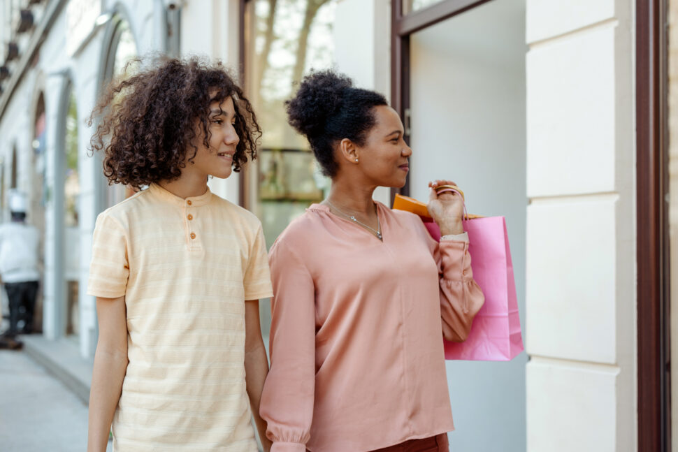 An African American Mother and Her Teenage Son Are Seen Walking Together in the City Center, Holding Shopping Bags, With Cheerful Expressions on Their Faces. The City Backdrop Features Modern Architecture, Bustling Streets, and Vibrant Storefronts, Creating an Energetic and Contemporary Atmosphere.