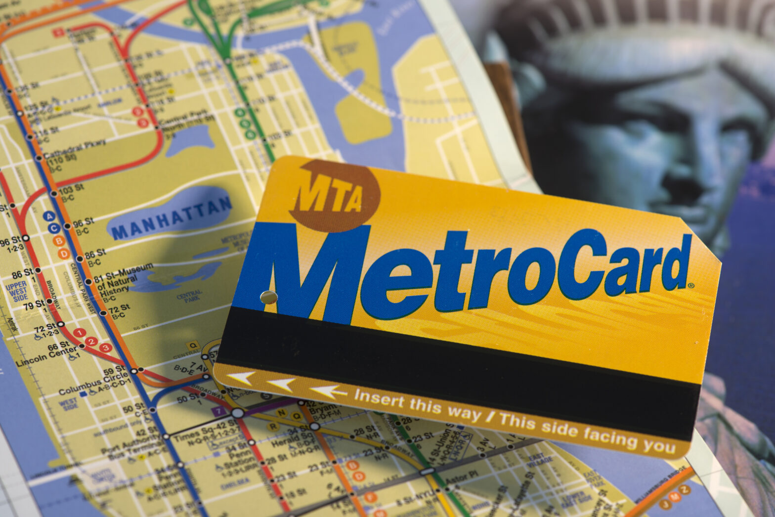 Front side of a standard rechargeable New York city MTA Metro Card, a NYC subway network map and a city travel guide in the background.