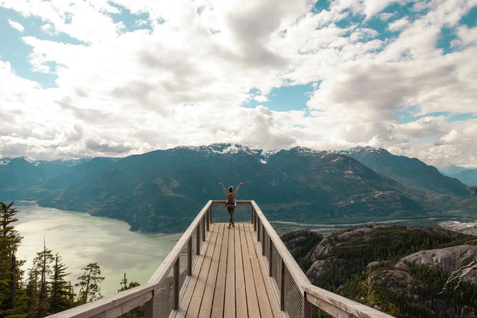 Person Standing With Hands on Air on Brown Wooden Dock With Overlooking View of Lake Under White Clouds and Blue Sky.