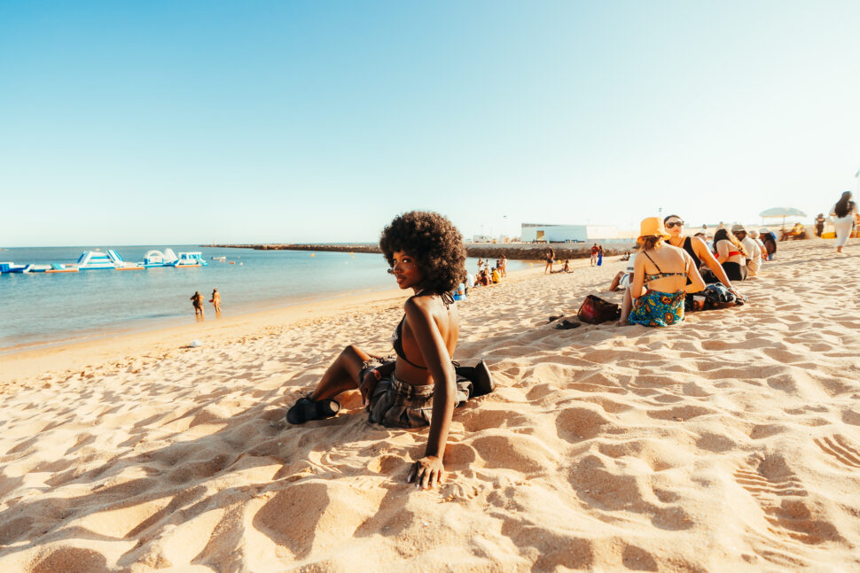 An attendee with an afro hairstyle sits on the sandy shore at Praia da Rocha, looking back toward the camera, with other festivalgoers relaxing nearby and inflatable structures floating in the calm blue water.