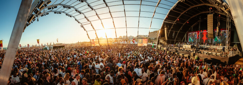 A massive crowd gathers under a large open-air canopy stage at Afro Nation Portugal during daylight, with the sun setting over the beach in the background.