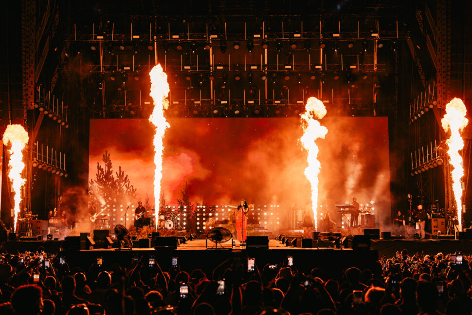 Tems sings center stage at Afro Nation Portugal as towering bursts of fire shoot upward, flanked by band members and a large illuminated backdrop.