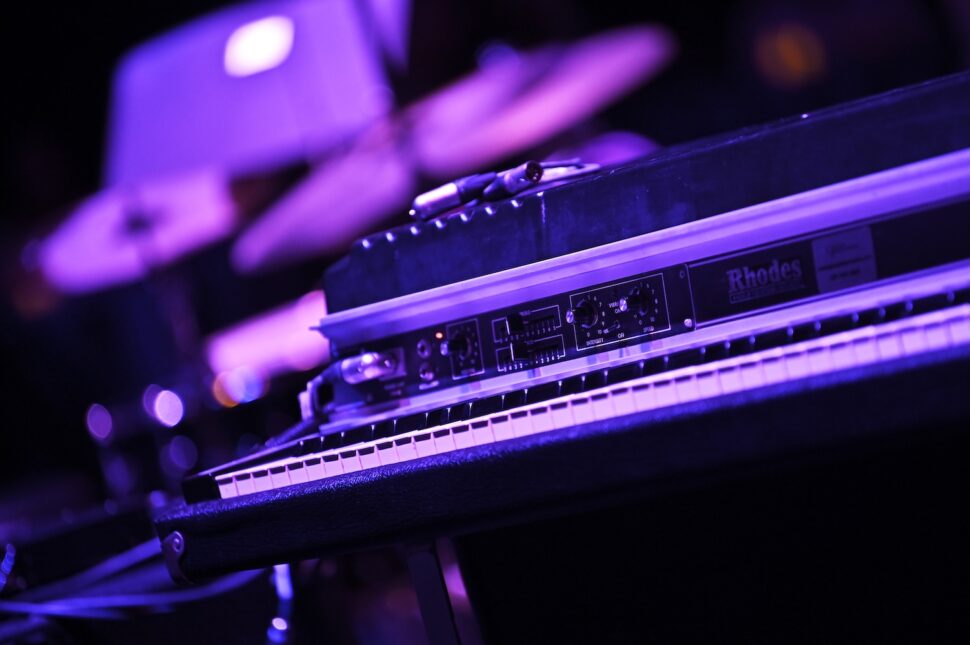 A close-up of a Rhodes electric piano illuminated by purple stage lights, with blurred drums and music gear in the background.